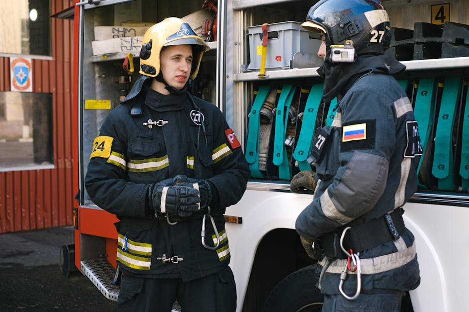 Two firefighters in full protective gear converse by a fire truck.